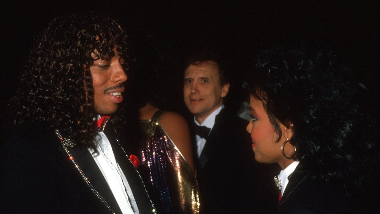Two people in formal attire, facing each other and smiling, are in the foreground at an event. A man in a tuxedo stands in the background. The lighting is dim and the mood appears festive.