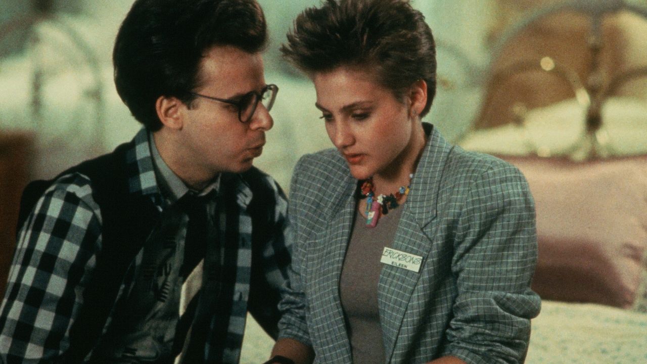 A man in glasses and a plaid shirt talks closely to a woman wearing a gray blazer and name tag. Both appear serious and are sitting together indoors with a blurred background.