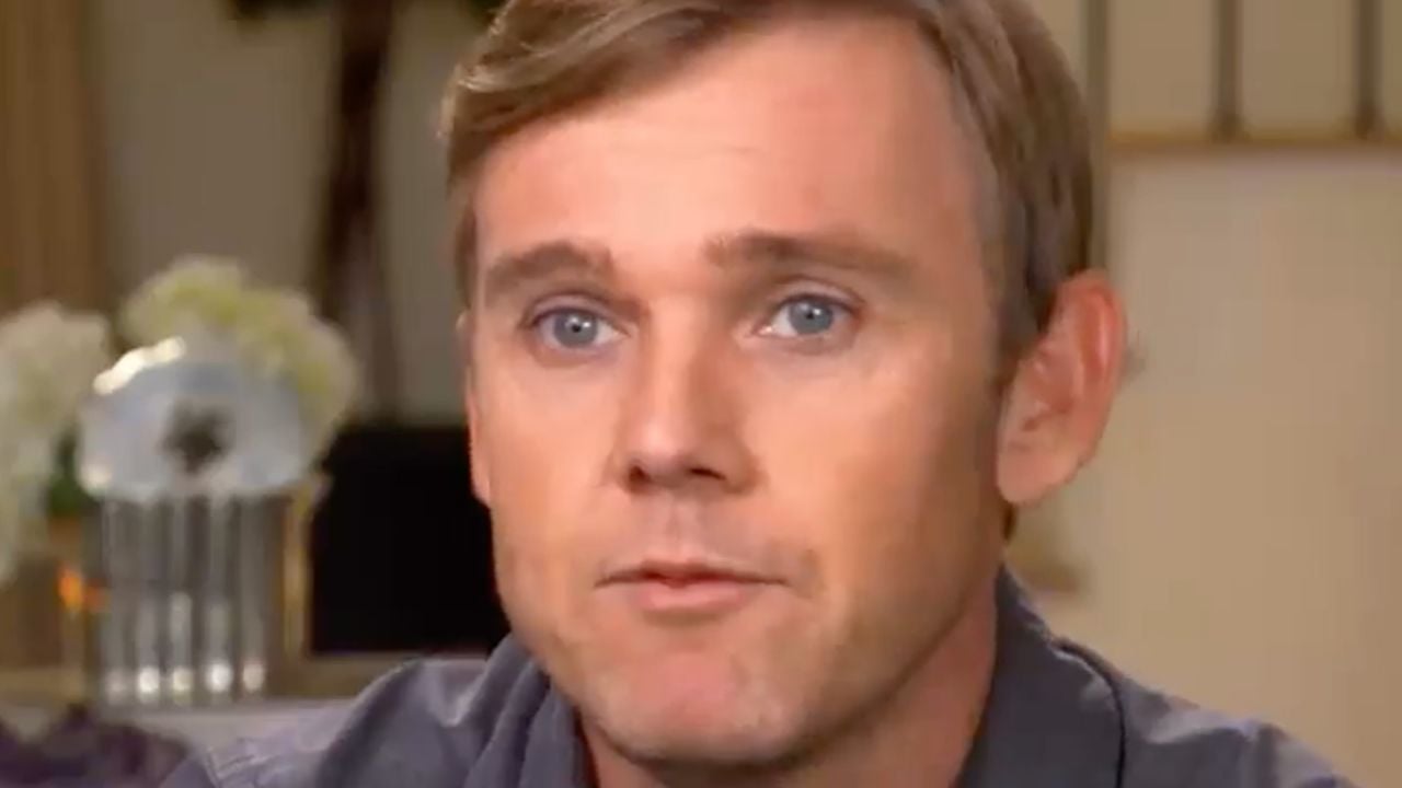 A man with light brown hair and blue eyes looks slightly past the camera. He is indoors, and there’s a blurred background with white flowers.
