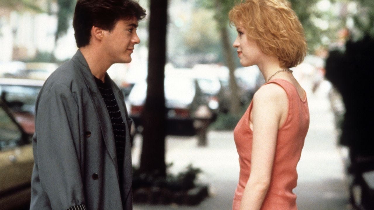 A young man and woman stand facing each other on a tree-lined street, having a conversation. The man wears a gray jacket and the woman wears a pink dress. Cars and greenery are visible in the background.