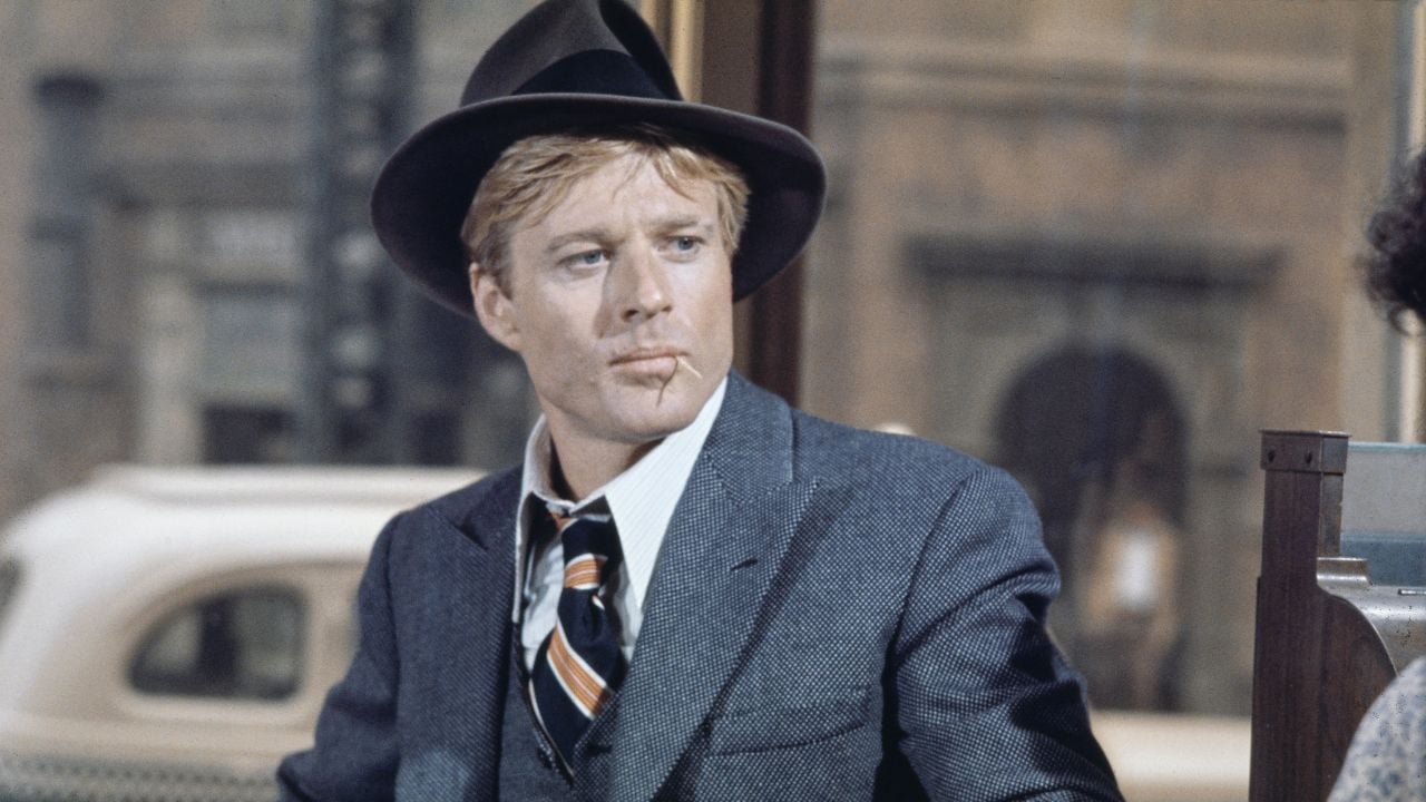 A man in a suit, striped tie, and fedora hat sits indoors. He has blonde hair and a serious expression. A blurred window view with an old car and building is in the background.