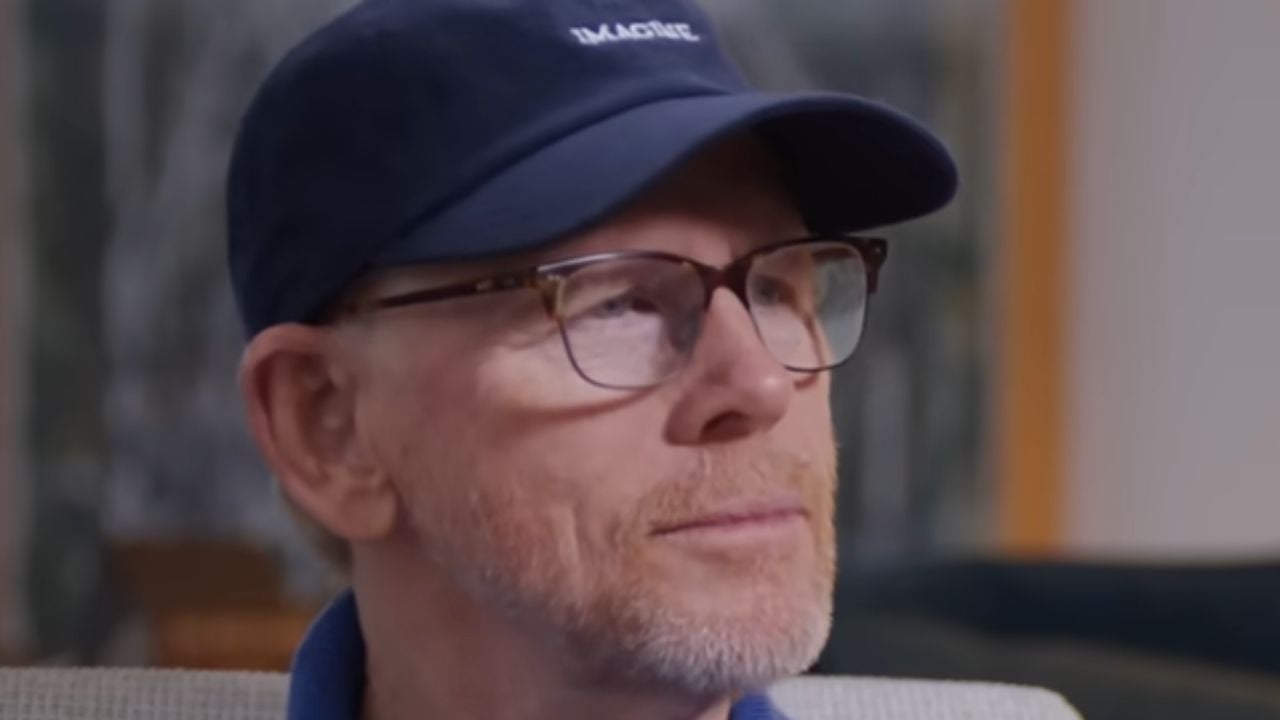 A man with light facial hair wears glasses and a dark blue cap that says “IMAGINE.” He is looking slightly to the side and appears thoughtful. The background is blurred.
