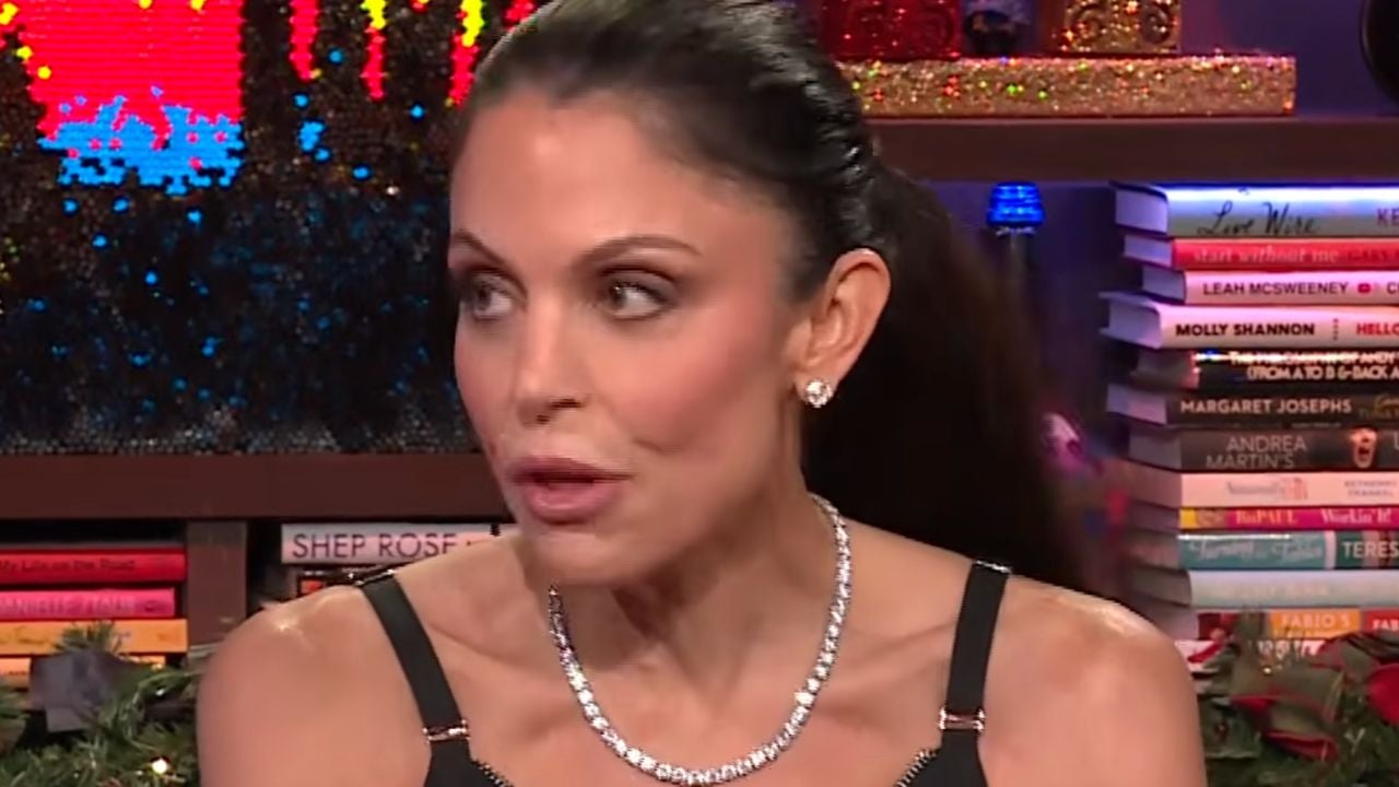 Woman with dark hair in a ponytail, wearing a black strappy top and a diamond necklace, sits in a colorful room with bookshelves in the background. She appears to be speaking or reacting to something.