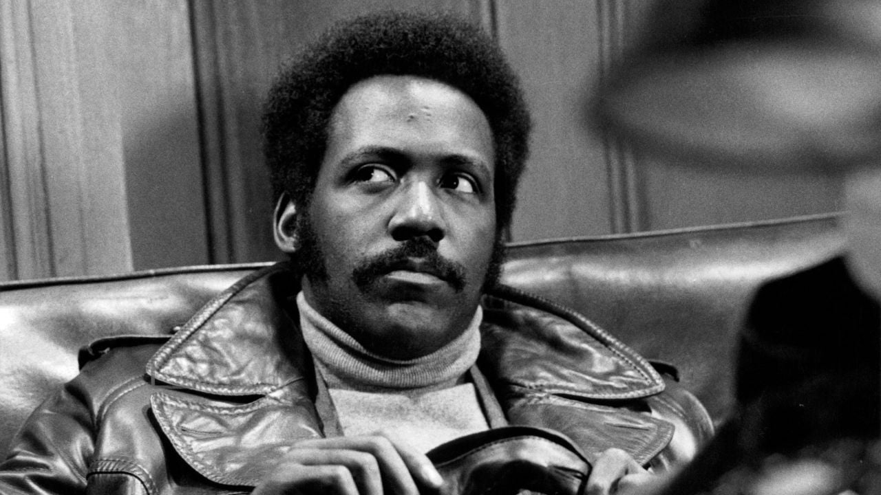 A man with an afro and mustache, wearing a leather jacket and turtleneck, sits on a couch and looks to the side with a serious expression. The background features wooden paneling.