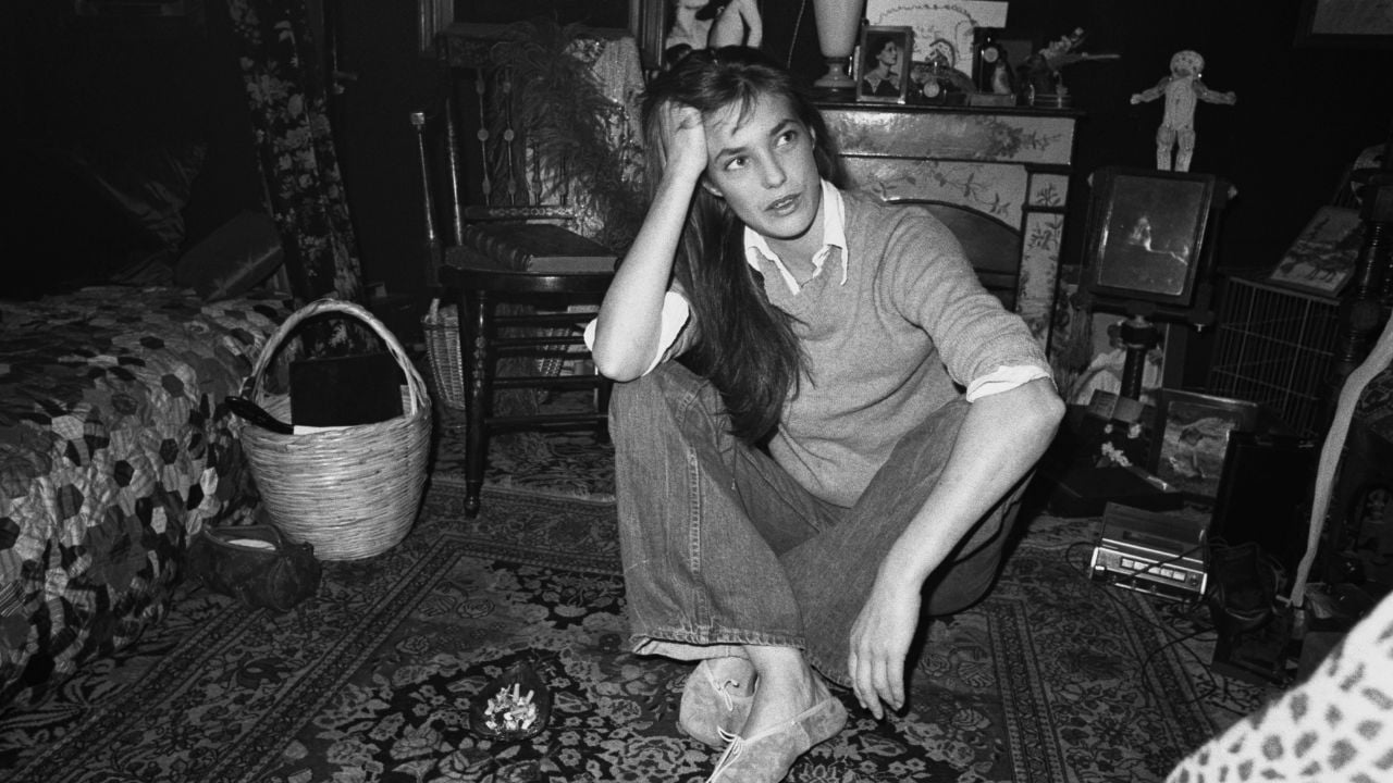 A woman sits cross-legged on a patterned rug in a cluttered room, resting her elbow on her knee and her head on her hand, gazing off thoughtfully. The space is filled with eclectic decor and personal items.
