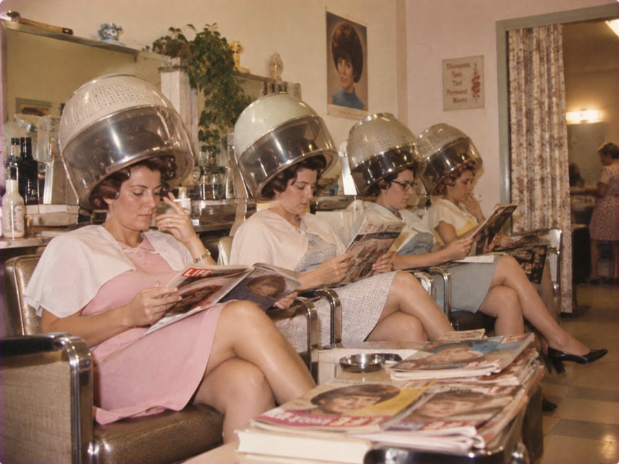 Four women sit under vintage hair dryers in a salon, reading magazines. A table in front holds more magazines. The salon has retro decor, mirrors, and posters on the walls.
