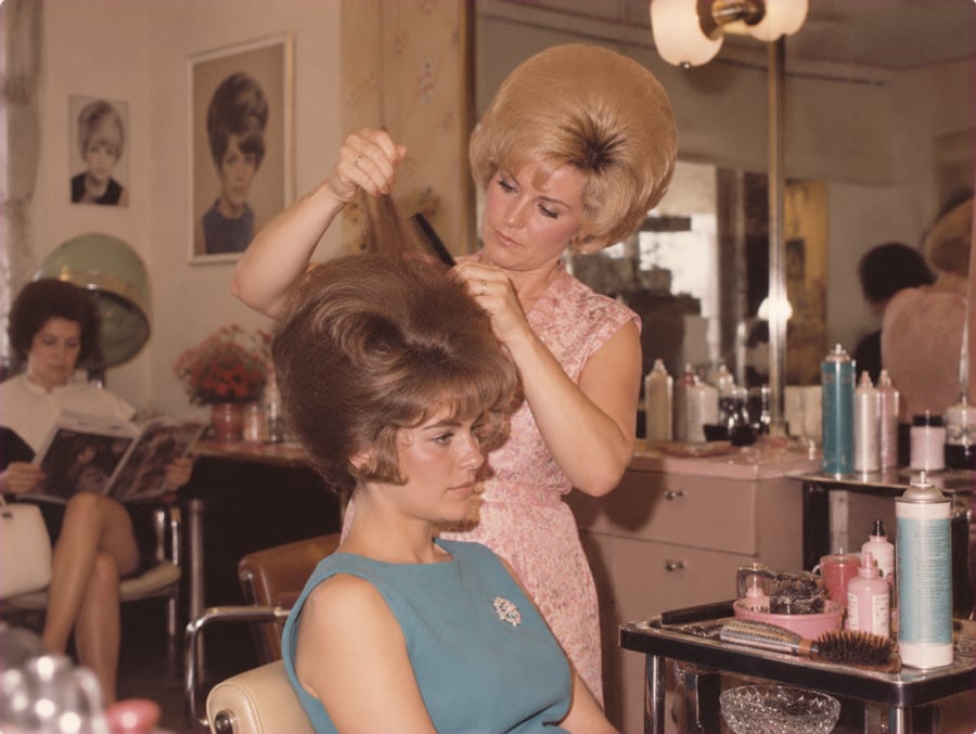 A hairdresser teases a woman’s hair into a beehive hairstyle in a vintage salon. Another woman reads a magazine in the background. The salon has retro décor and various hair products on the counter.