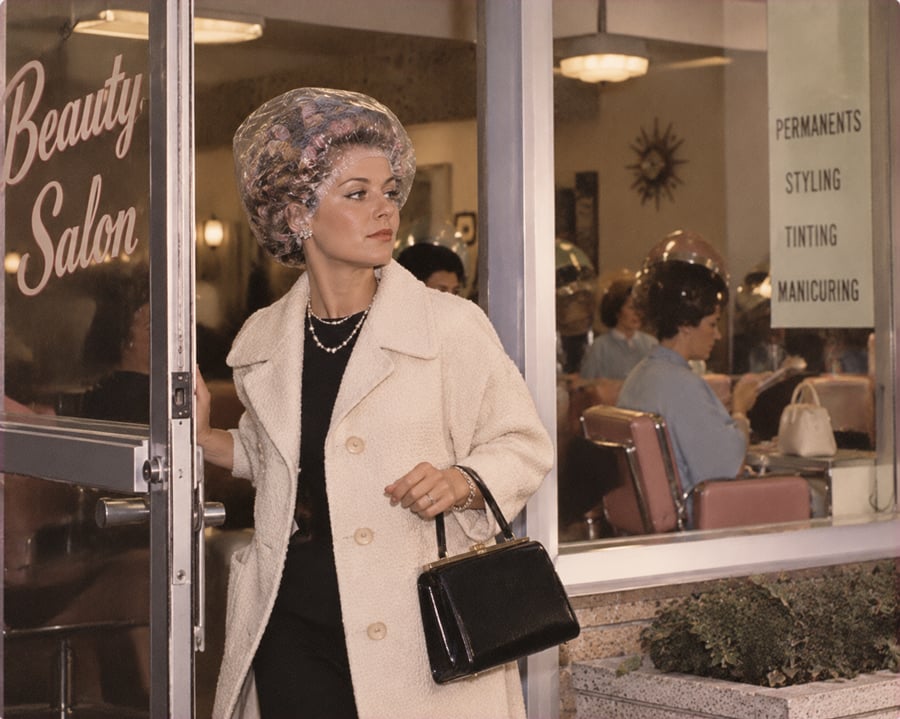 A woman in a cream coat and hair curlers exits a beauty salon, holding a black handbag. Inside, several women sit under hair dryers. A sign on the window lists salon services: permanents, styling, tinting, manicuring.