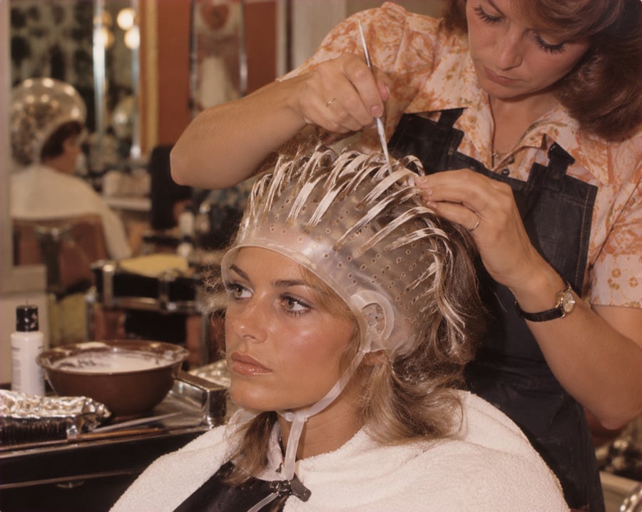 A woman sits in a salon chair wearing a highlighting cap while a stylist pulls strands of her hair through the holes and applies hair dye with a brush.