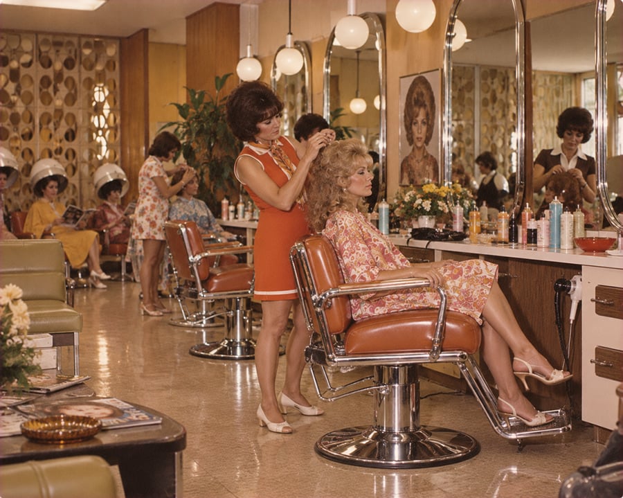 A hairstylist in an orange dress styles a seated woman’s hair in a retro salon, with other women under dryers in the background; mirrors, products, and vintage decor fill the space.