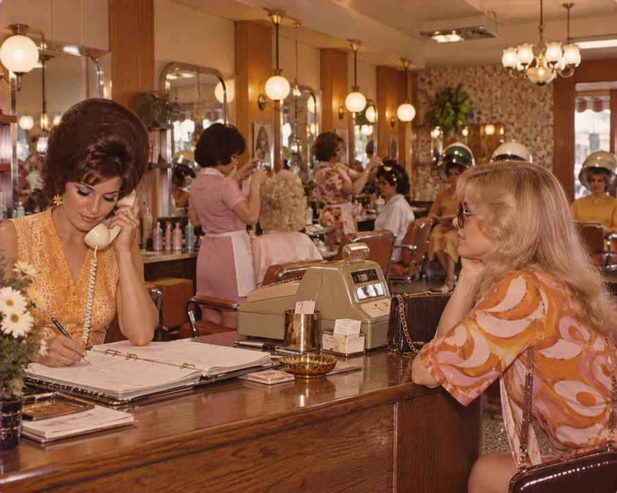 Two women sit at a salon reception desk. One is on the phone, writing in a large book, while the other waits. Several women are visible in the background getting their hair styled. The scene looks vintage, likely from the 1970s.