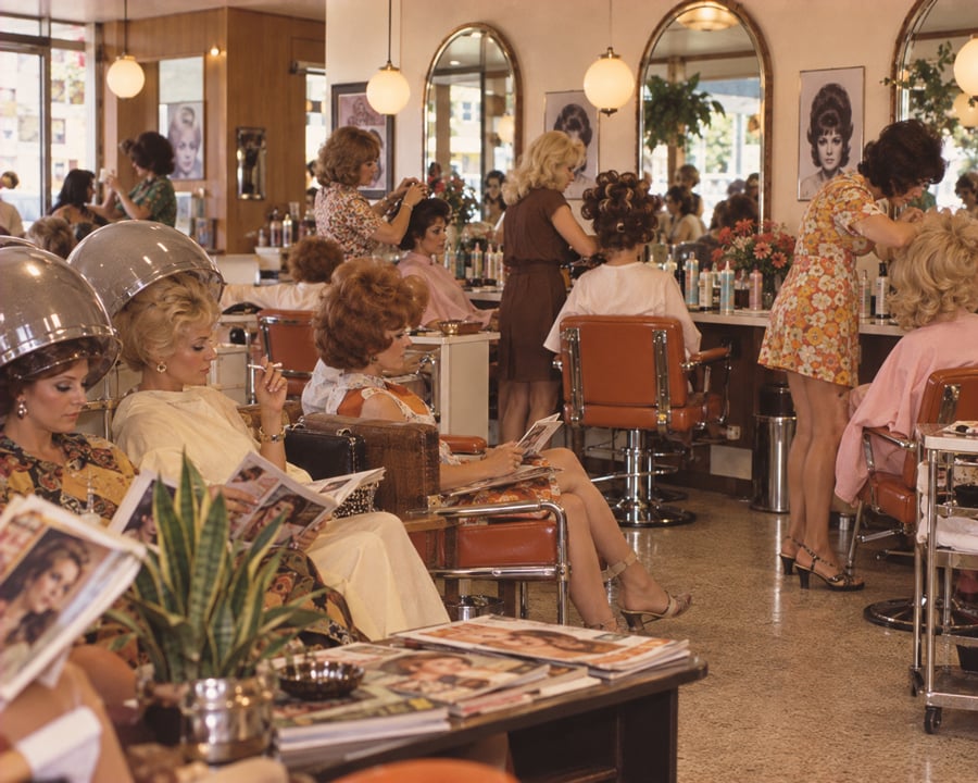 Women sit under hair dryers and read magazines while others have their hair styled in a busy, vintage beauty salon with retro decor, orange chairs, mirrors, and hair product bottles on counters.