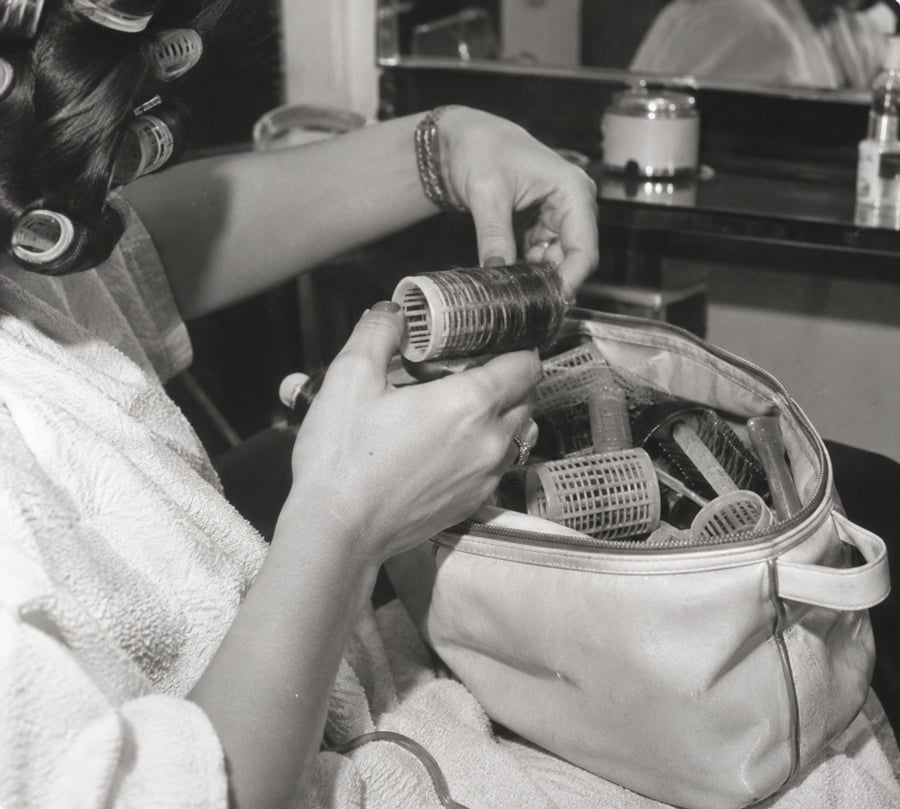 A person wearing hair rollers holds another roller above an open bag filled with more rollers, sitting at a vanity with a towel over their shoulders.