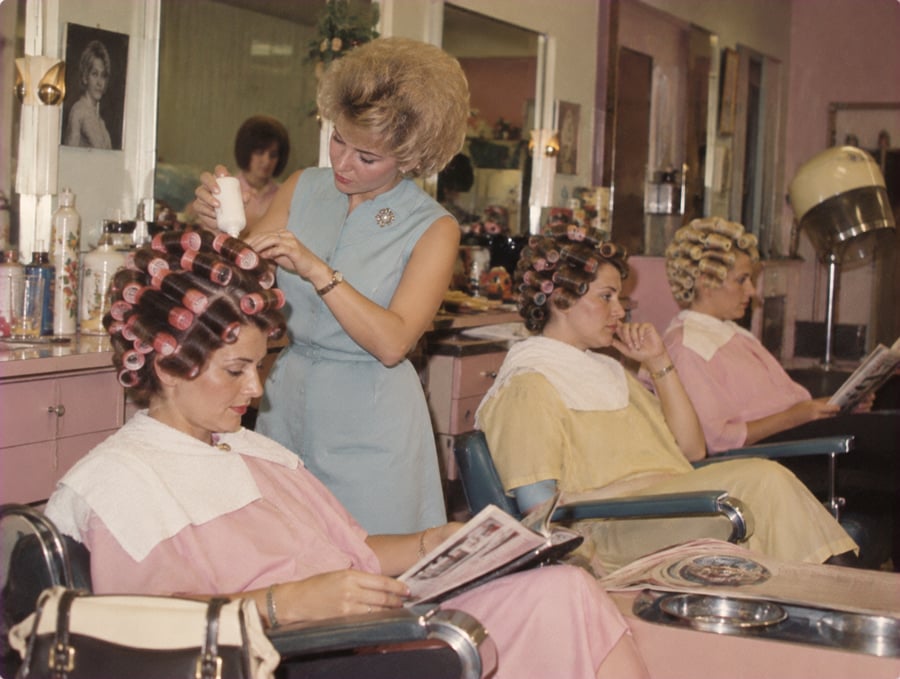 Three women sit in salon chairs with curlers in their hair and capes on, while a stylist applies product to one client. Two clients read magazines. The salon has pink walls, mirrors, and various hair products on counters.