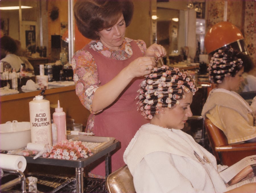 A hairdresser applies curlers to a woman’s hair at a salon. The woman wears a salon cape and reads a magazine. Bottles labeled “Acid Perm Solution” are on the counter. Other clients are visible in the background.