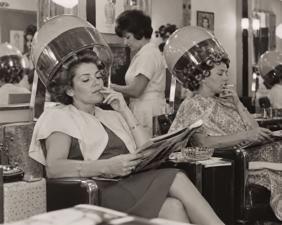 Two women sit under vintage hair dryers at a salon, reading magazines and smoking. A hairstylist works in the background. The scene appears to be from the mid-20th century.