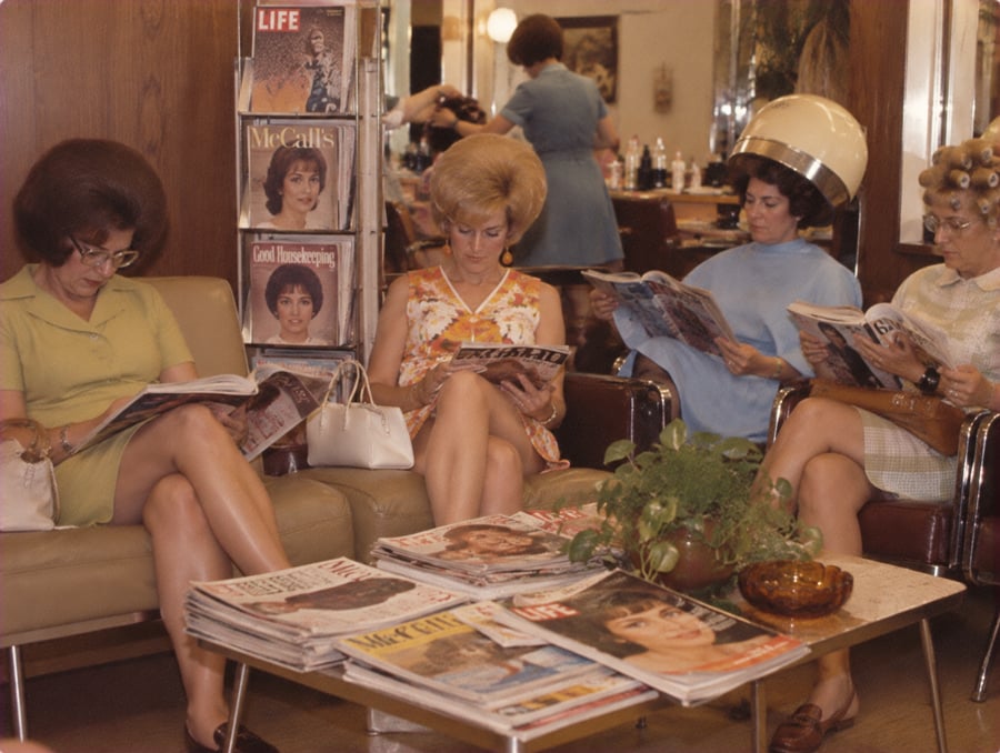 Four women sit in a vintage beauty salon, reading magazines. Some have hair in rollers or under dryers. A rack holds magazines like LIFE and McCall’s. A table with magazines and a potted plant is in front of them.