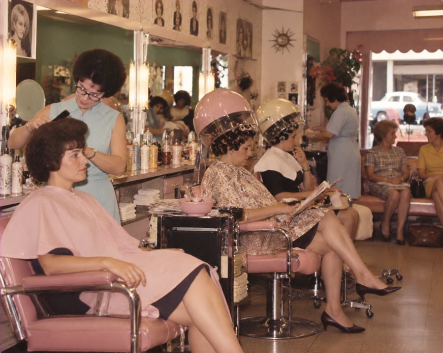 Women sit in pink chairs at a busy vintage hair salon. Some have hair dryers over their heads, while a stylist works on a client’s hair. Magazines and hair products fill the vibrant, retro-styled room.