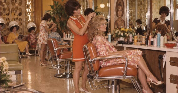 A woman in an orange dress styles another woman’s hair in a vintage salon. Other women sit under hair dryers. The room has mirrors, beauty products, and retro decor.