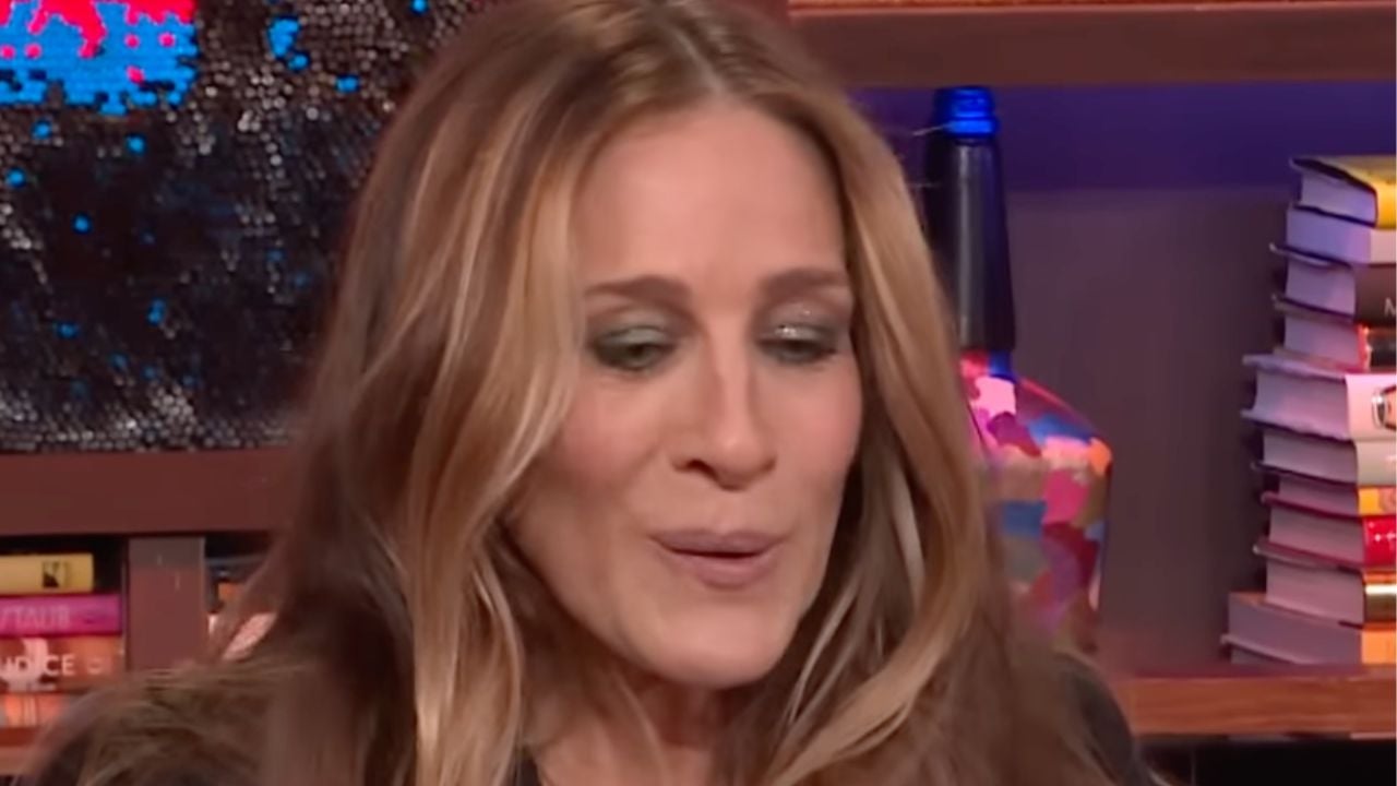 A woman with long, wavy blonde hair and shimmery eye makeup is looking downward. Books and a colorful lamp are visible in the background on shelves.
