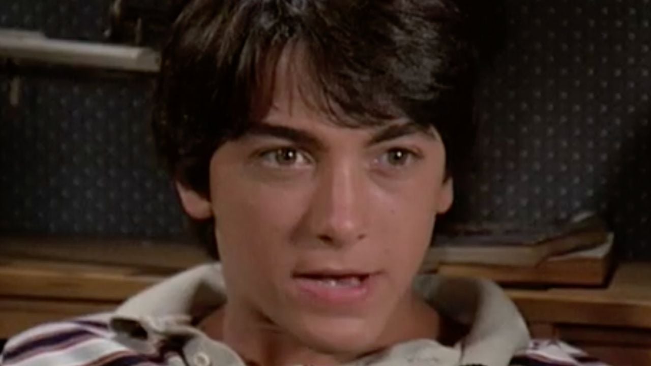 A young person with dark hair and striped shirt looks ahead with a serious expression. A book and a patterned background are visible behind them.