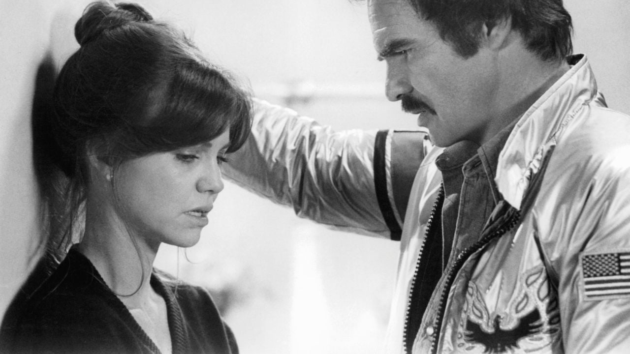 A woman with dark hair looks down sadly as a man in a shiny jacket with an eagle patch leans against the wall above her, looking at her seriously. The scene is in black and white.