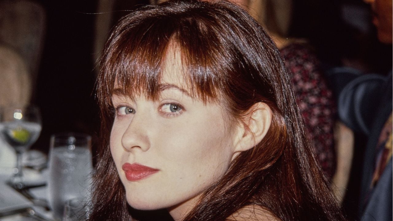A woman with long brown hair and bangs looks over her shoulder at the camera while sitting at a table set with glasses and plates in a restaurant.