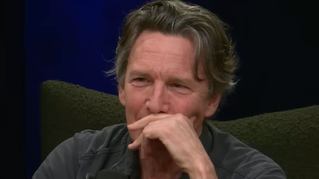 A man with gray hair sits in a dark room, resting his hand over his mouth while looking thoughtful. He wears a dark shirt and is seated on a textured dark chair.