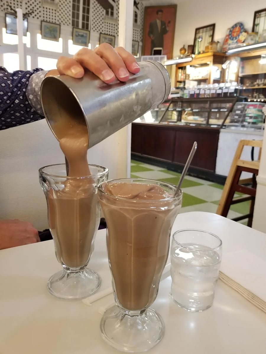A hand pours a thick chocolate milkshake from a metal shaker into a glass beside another filled glass, with a spoon and a glass of water on a café table.