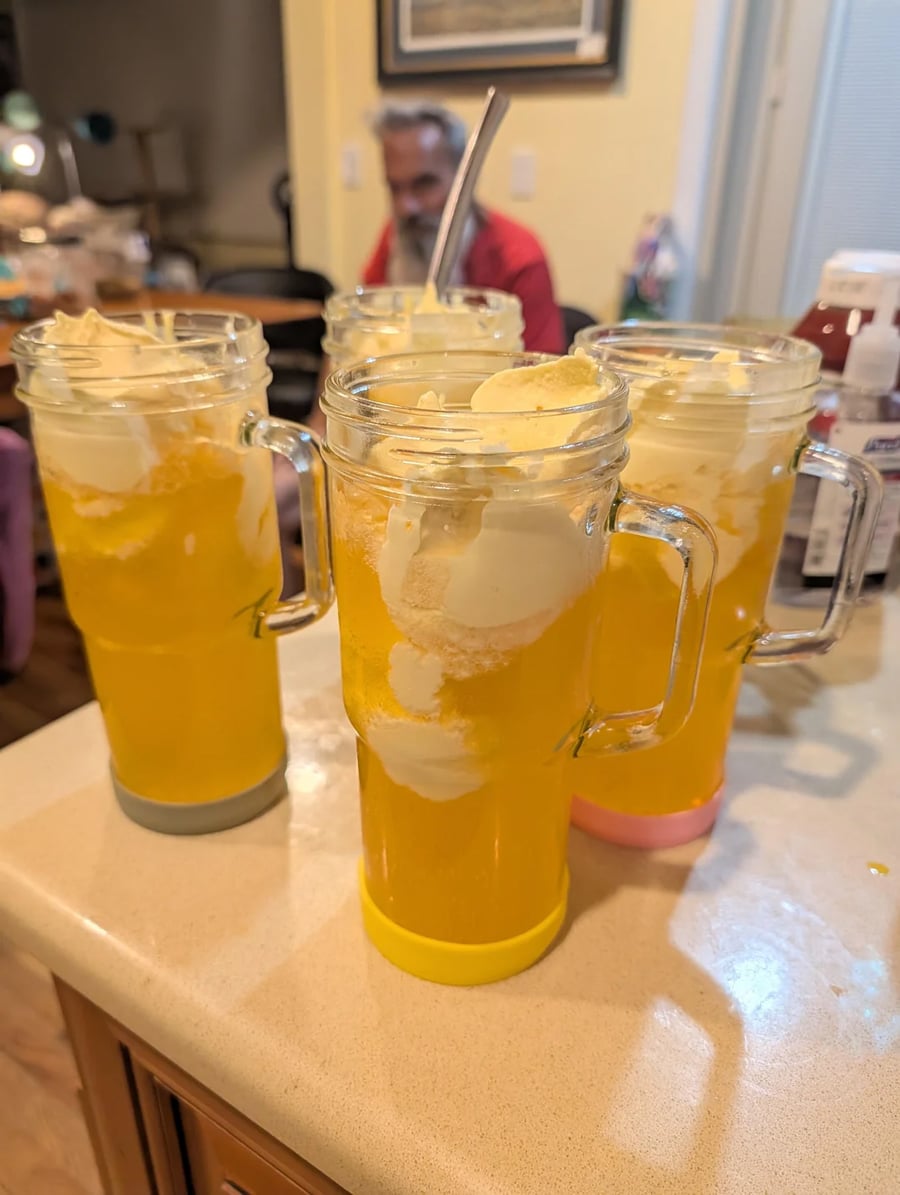 Four tall mugs filled with yellow soda and scoops of vanilla ice cream sit on a kitchen counter. A person is visible in the background, and kitchen items are scattered nearby.
