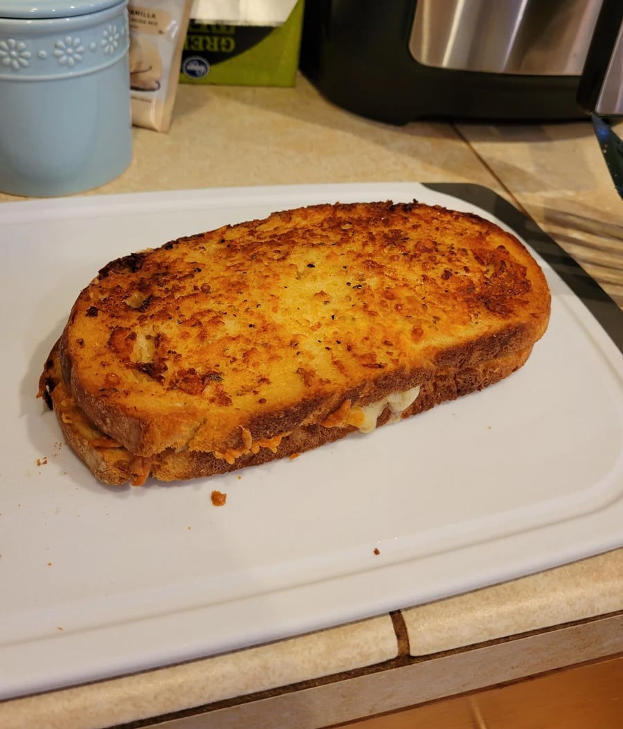 A golden brown grilled cheese sandwich with melted cheese peeking out, sitting on a white cutting board on a kitchen counter.
