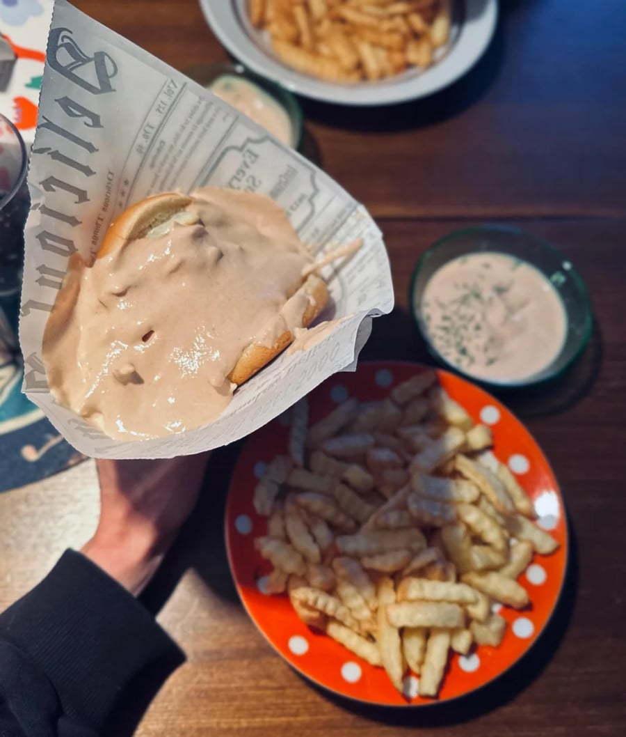 A hand holds a burger wrapped in newspaper print paper and covered with sauce. Below, there’s a red plate with crinkle-cut fries and a small bowl of dipping sauce on a wooden table.