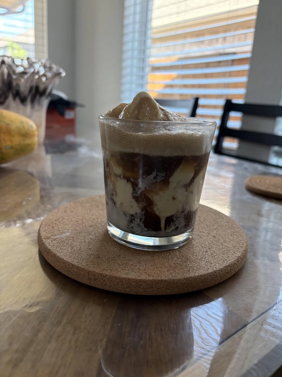 A glass of iced coffee with cream sits on a round cork coaster atop a wooden table, with sunlight streaming through window blinds in the background.