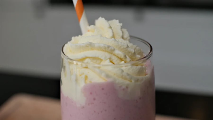 A close-up of a pink milkshake in a clear glass, topped with swirls of whipped cream and an orange striped straw.