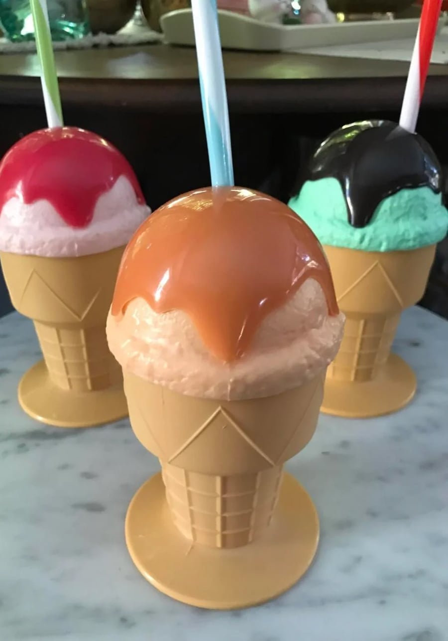 Three plastic ice cream cone cups with fake ice cream tops in red, caramel, and mint colors, each with a straw sticking out, placed on a marble surface.