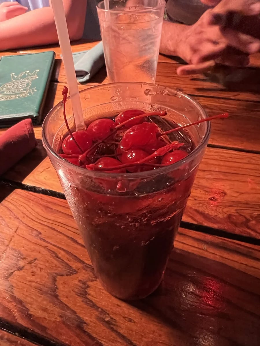 A glass of dark soda topped with many maraschino cherries and a straw sits on a wooden table in a restaurant, with menus and people’s hands visible in the background.