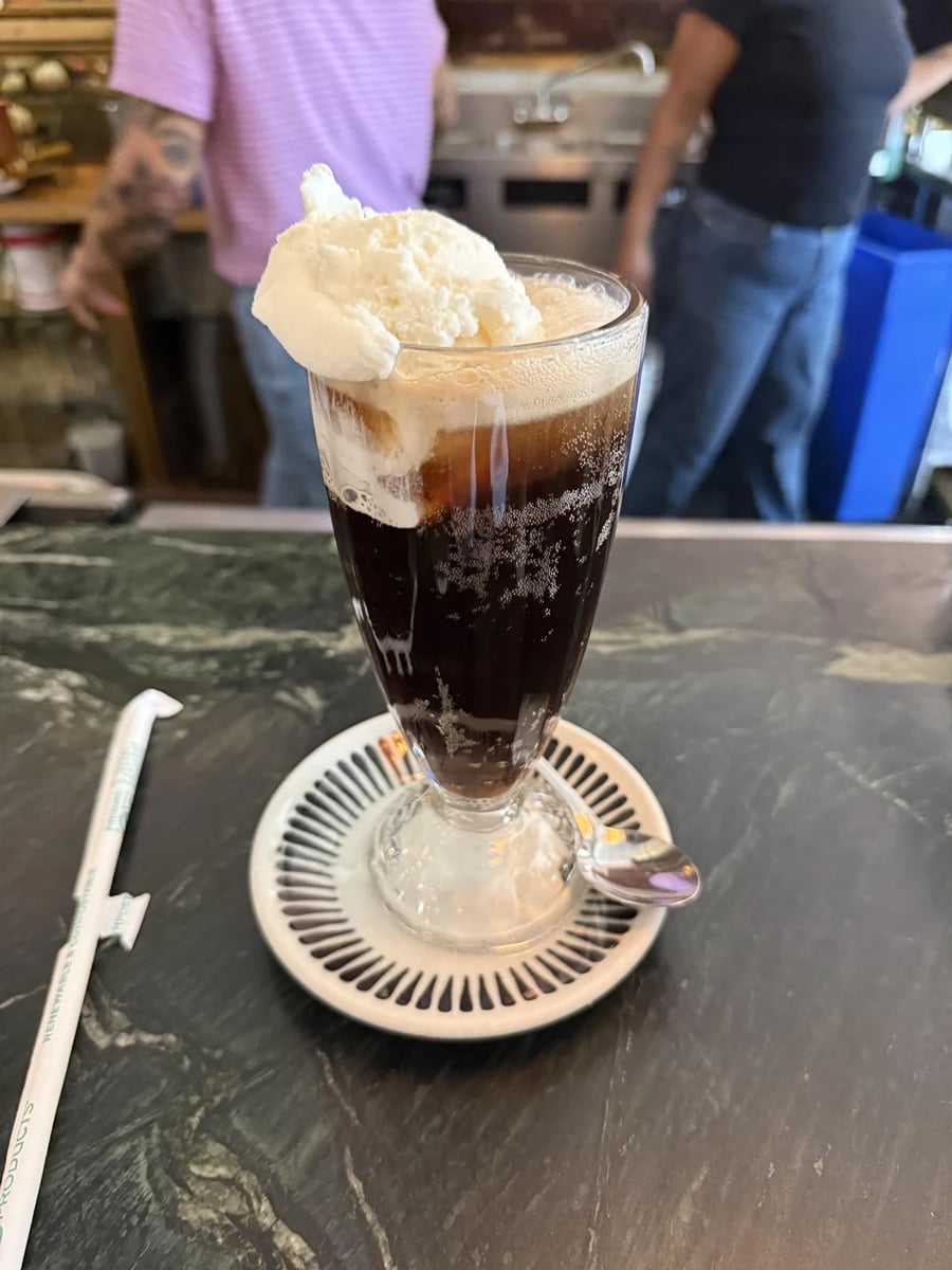 A glass of iced coffee with a scoop of vanilla ice cream on top, served on a saucer with a small spoon, sits on a marble counter. Two people are blurred in the background.