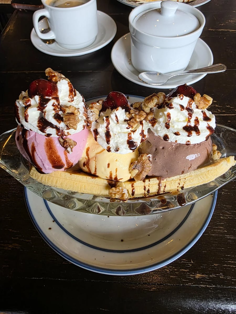 A banana split with three scoops of ice cream—strawberry, vanilla, and chocolate—topped with whipped cream, chocolate syrup, walnuts, and cherries, served in a glass dish on a plate. Coffee cups are in the background.