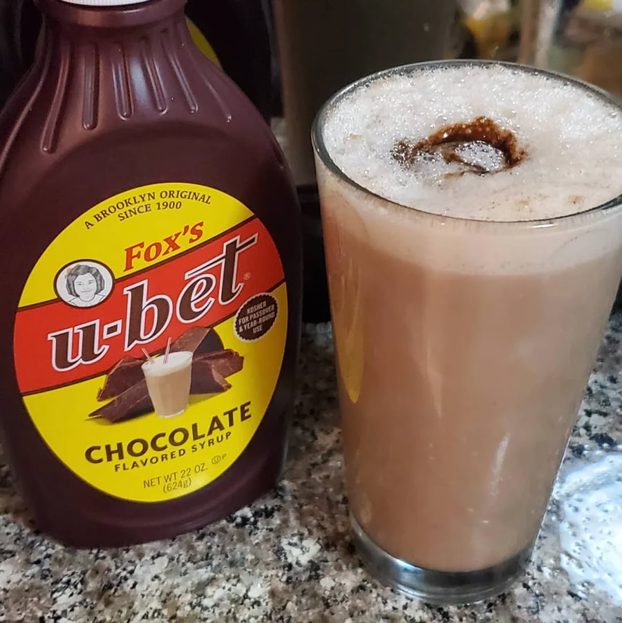 A bottle of Fox’s U-Bet chocolate syrup stands next to a glass of frothy chocolate milk topped with syrup, on a speckled kitchen countertop.