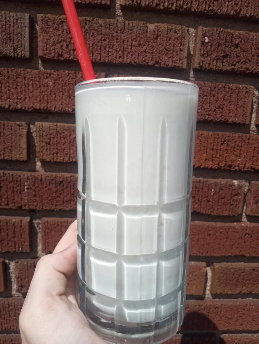 A hand holds a tall glass of a creamy white milkshake with a red straw in it, set against a brick wall in bright sunlight.