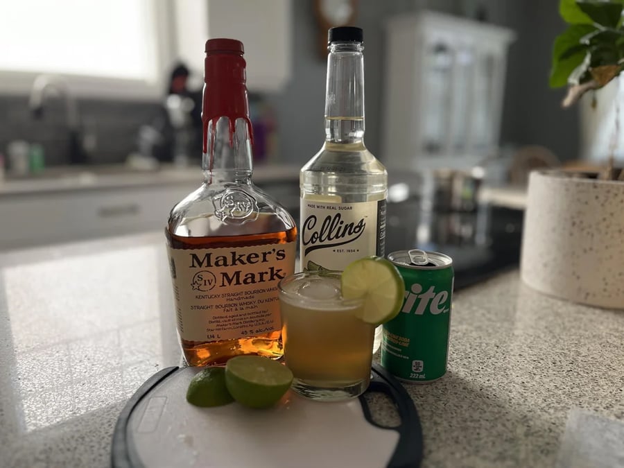 A bottle of Maker’s Mark whiskey, Collins mixer, a can of Sprite, a glass with a drink, and sliced limes are arranged on a kitchen countertop.