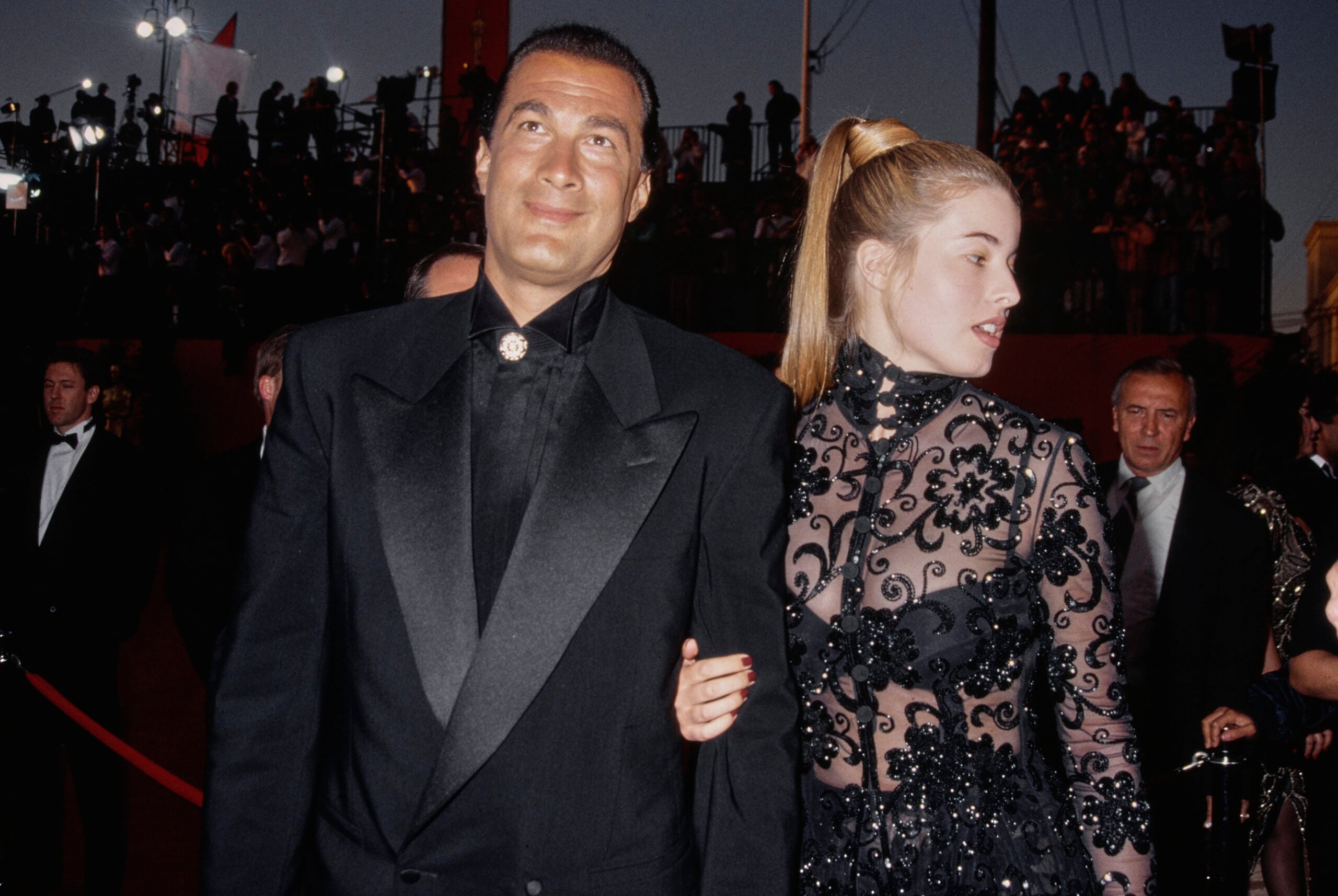 A man in a black suit and a woman in a sheer, black lace dress walk together on a red carpet at an evening event, with crowds and photographers in the background.