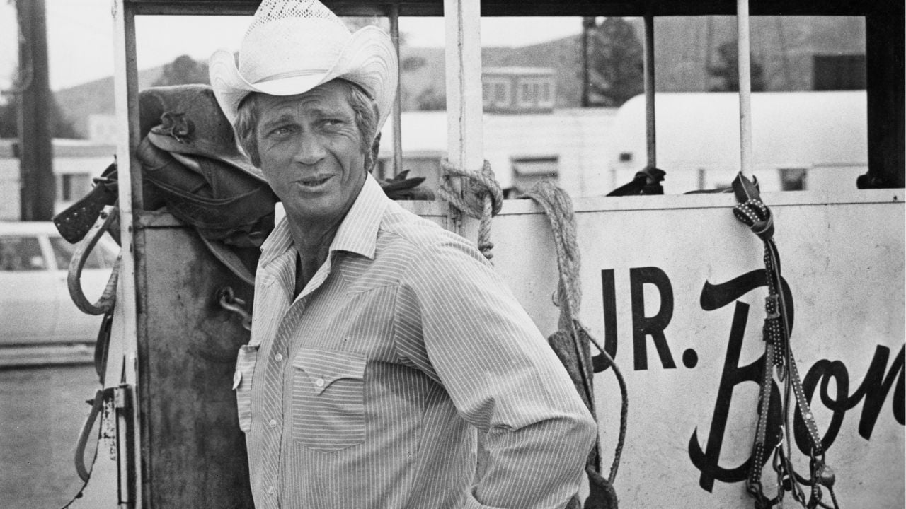 A man in a white cowboy hat and striped shirt stands beside a truck with saddles and a sign reading “JR BONE.” He looks to the side with a serious expression.