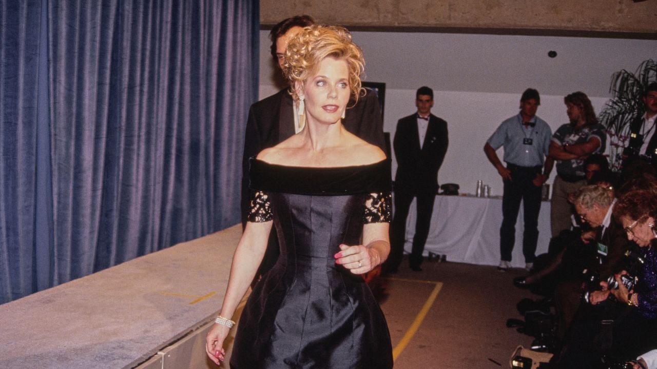 A woman in an elegant black off-the-shoulder gown walks indoors, with people seated and standing around her. She has blonde hair styled up and wears long earrings. The setting appears to be a formal event.