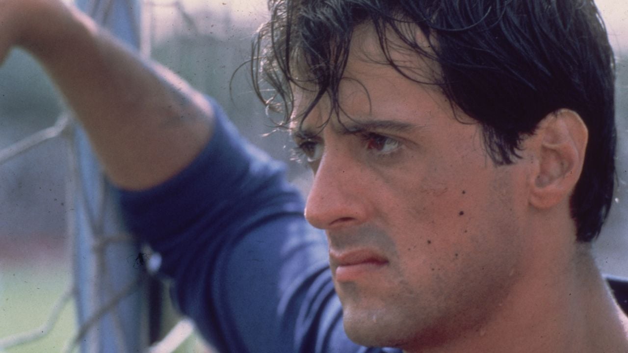A man with dark hair and a serious expression looks through a chain-link fence, wearing a blue shirt. His face is sweaty and he appears deep in thought.