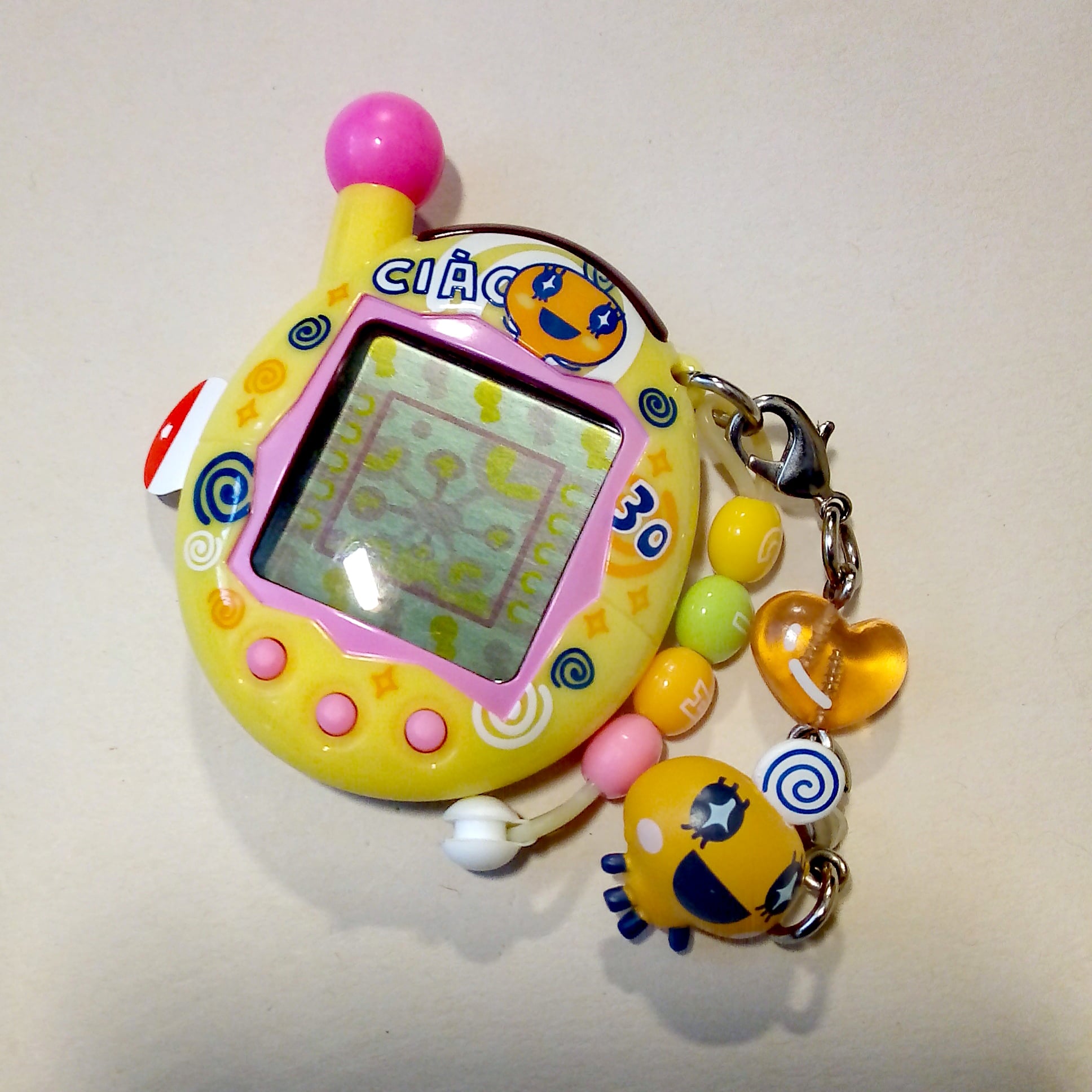 A yellow Tamagotchi toy with pink buttons, a colorful screen, and character-themed charms, including beads and a small orange heart, attached by a keychain.