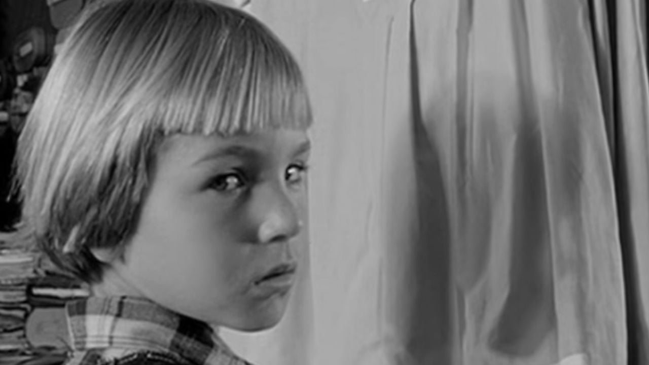 A young child with straight, light-colored hair and bangs looks seriously over their shoulder, wearing a plaid shirt. The background is blurred with indistinct shapes and a curtain. The image is in black and white.