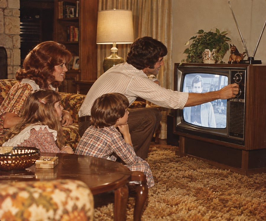 A family sits in a living room watching a black-and-white TV. The father adjusts the TV dial while the mother and two children sit nearby on a patterned carpet, all focused on the screen.
