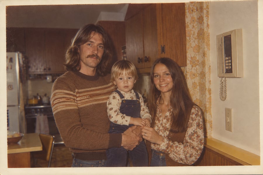 A man, woman, and young child pose in a vintage kitchen. The man has long hair and a mustache, holding the child. The woman stands beside them, all smiling. The kitchen has wood cabinets and a wall-mounted rotary phone.