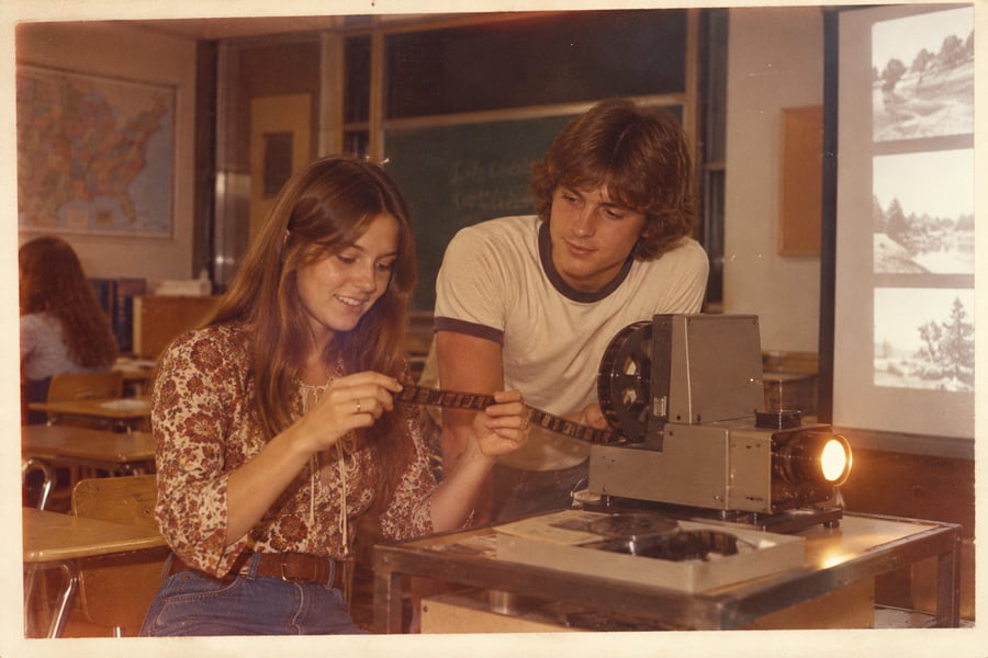 Two students in a classroom examine a film strip by a projector. The girl holds the film while the boy looks on. A map and desks are visible in the background, with images projected on the wall.